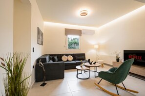Una sala moderna con un sofá, dos mesitas auxiliares, un sillón mecedor verde, un televisor y una ventana con persianas.