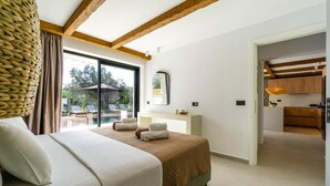 Ein Schlafzimmer mit einem großen Bett, einem Fenster mit Blick auf Gartenmöbel, einer Holzbalkendecke und einer durch eine offene Tür sichtbaren Küchenzeile.