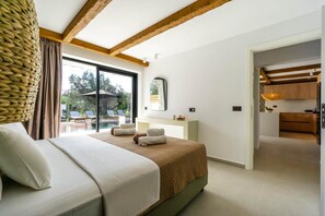 Ein Schlafzimmer mit einem großen Bett, einem Fenster mit Blick auf Gartenmöbel, einer Holzbalkendecke und einer durch eine offene Tür sichtbaren Küchenzeile.