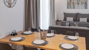 A dining area with a wooden table set for four, a sofa in the background, and a wall clock. - Zoelgrek Luxury Suite 2 Apartment with Shared Terrace, Wi-Fi & Air Conditioning (Iraklio)
