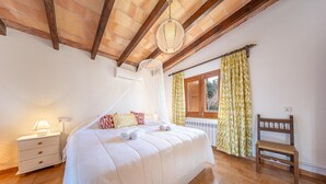 A bedroom with a bed, bedside table, lamp, window with curtains, and a wooden bench. - Villa del la Vilar in Mallorca (Pollença)