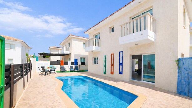 A swimming pool with a tiled deck, a covered outdoor seating area, and a building with balconies and doors. - Villa Chromata in Pernera (Paralimni)