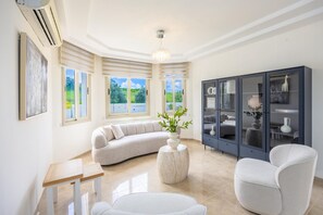 A modern living room with a curved sofa, a round table, a cabinet with glass doors, and a ceiling fan. - Villa Lefkos in Argaka (Argaka)