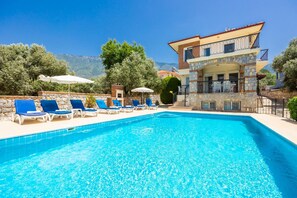 A pool with blue water, surrounded by lounge chairs and umbrellas, with a building featuring balconies and stone walls in the background. - Golden Trio Villa in Oludeniz (Fethiye)