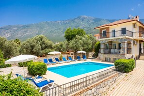 A pool with lounge chairs, a stone building with balconies, and a mountainous backdrop. - Golden Trio Villa in Oludeniz (Fethiye)
