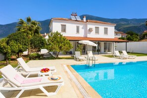 Eine weiße Villa mit Swimmingpool, Liegestühlen und Bergblick.