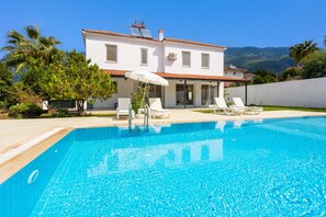 Eine weiße Villa mit Swimmingpool, Liegestühlen und Bergblick.