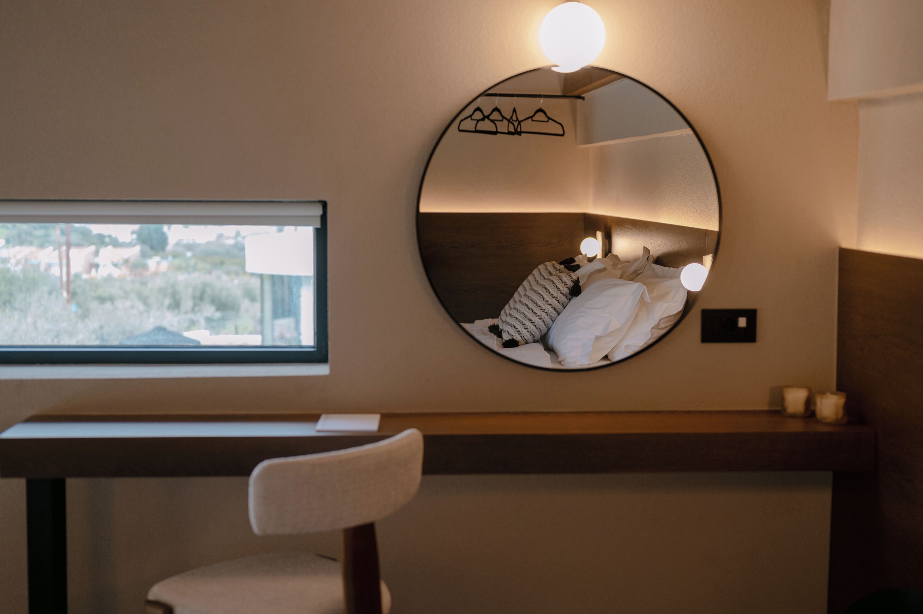 A bedroom with a round mirror, bedside table, chair, and a window with a view.