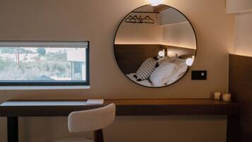 A bedroom with a round mirror, bedside table, chair, and a window with a view.