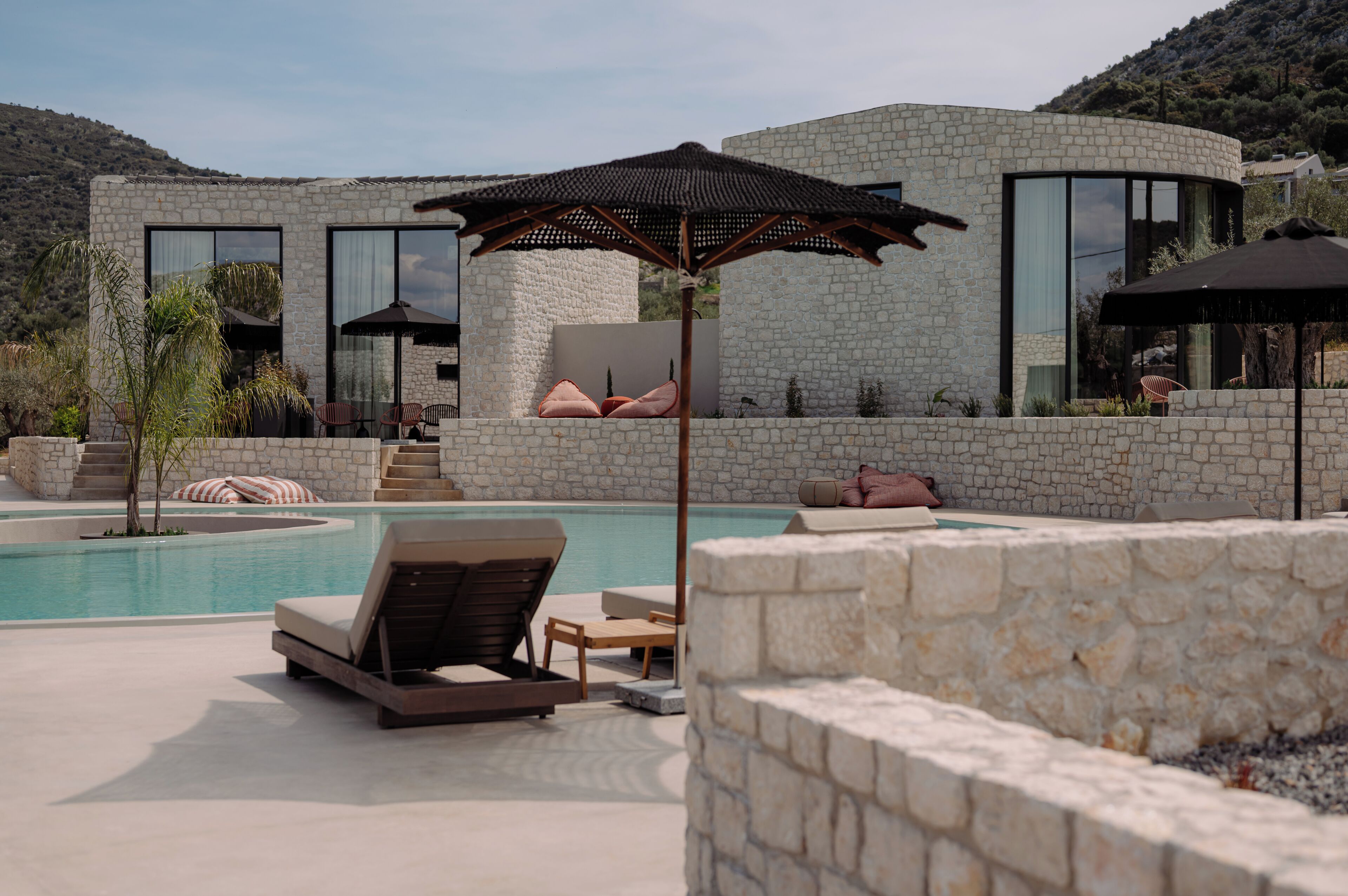 A modern poolside area with lounge chairs, umbrellas, and a stone building with large windows.
