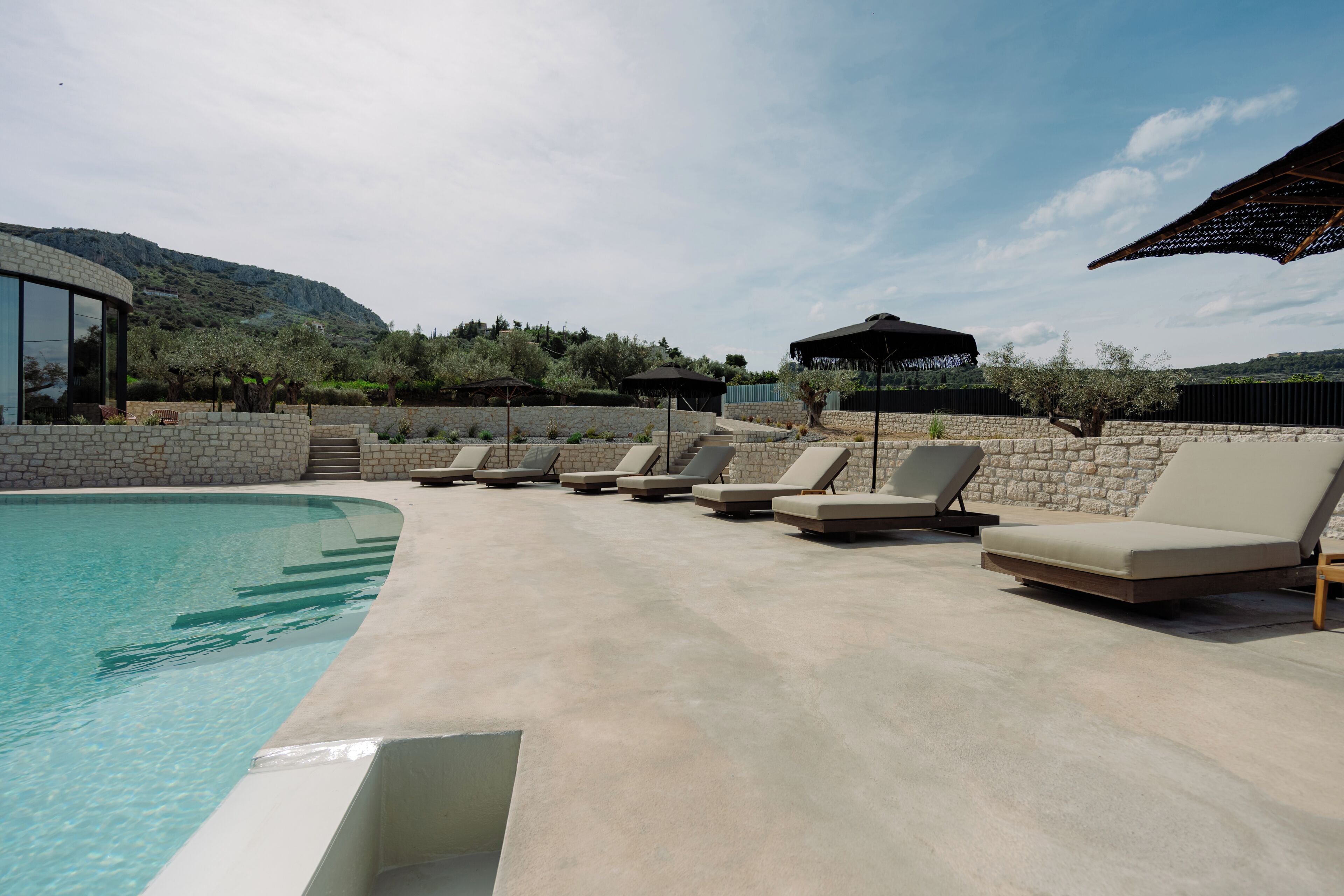 A pool area with lounge chairs and umbrellas, surrounded by stone walls and a view of hills.