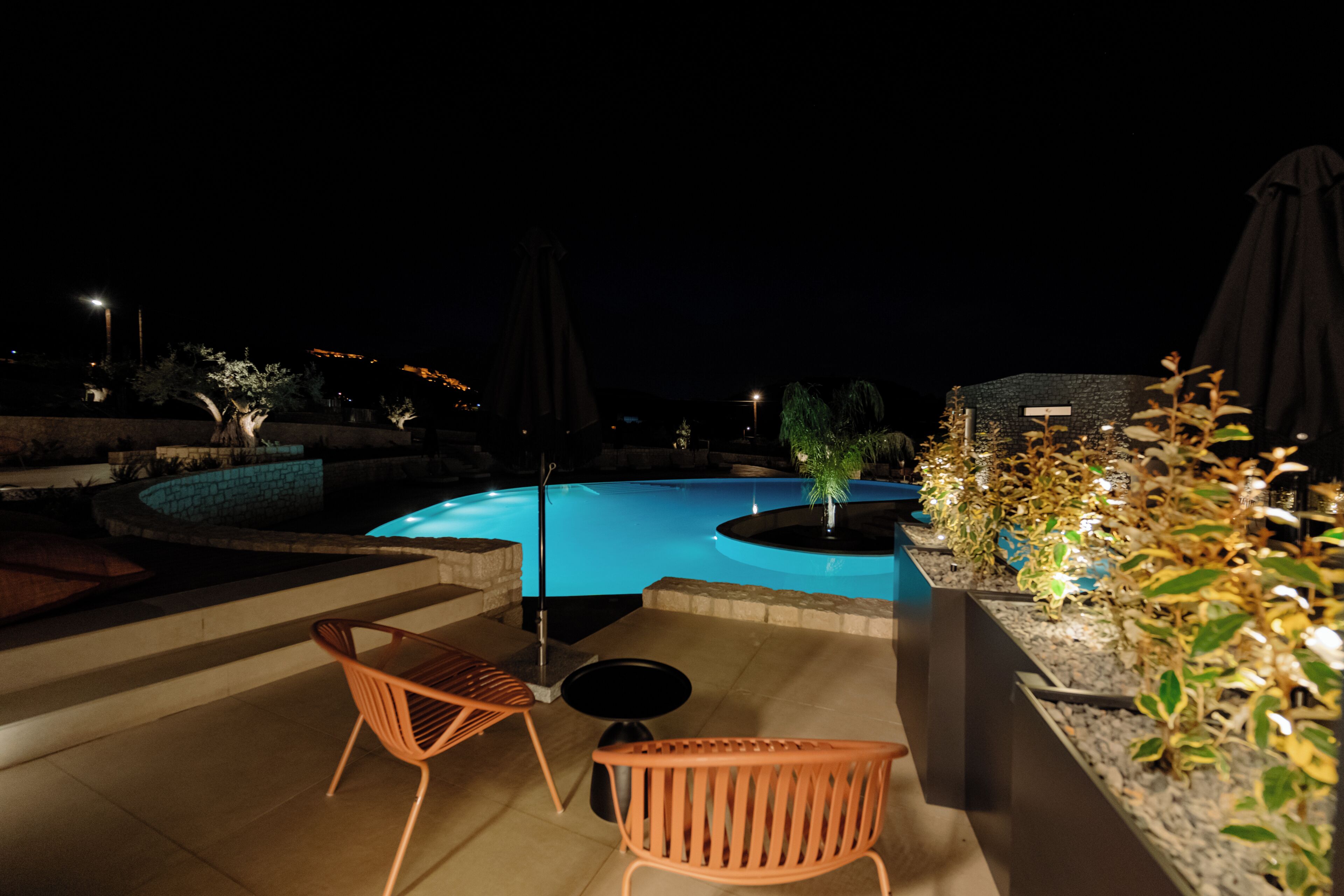 A nighttime pool area with a curved pool, two chairs, and a small table.