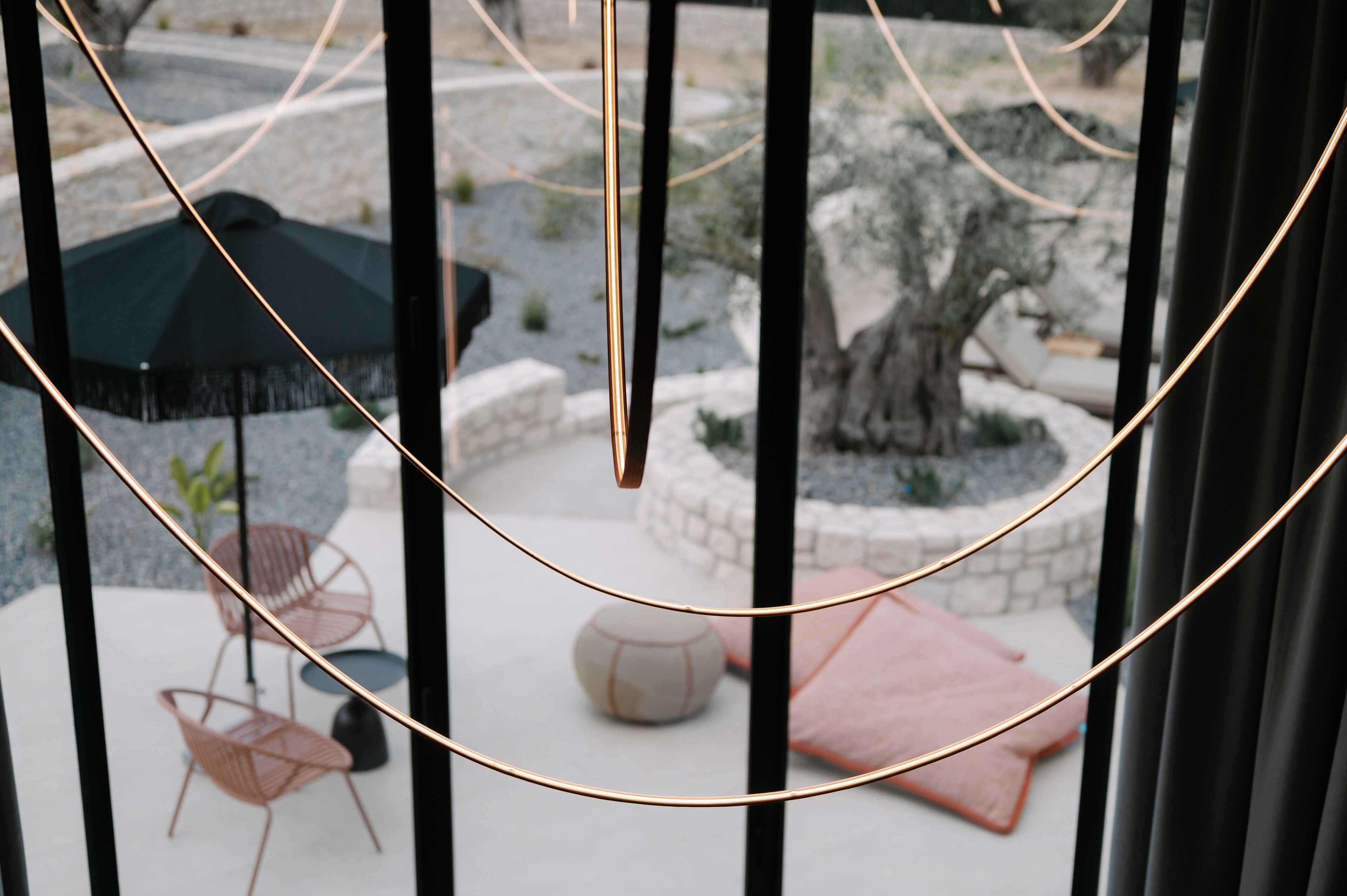 A view through a window with a black metal frame, a black umbrella, and a round table with chairs.