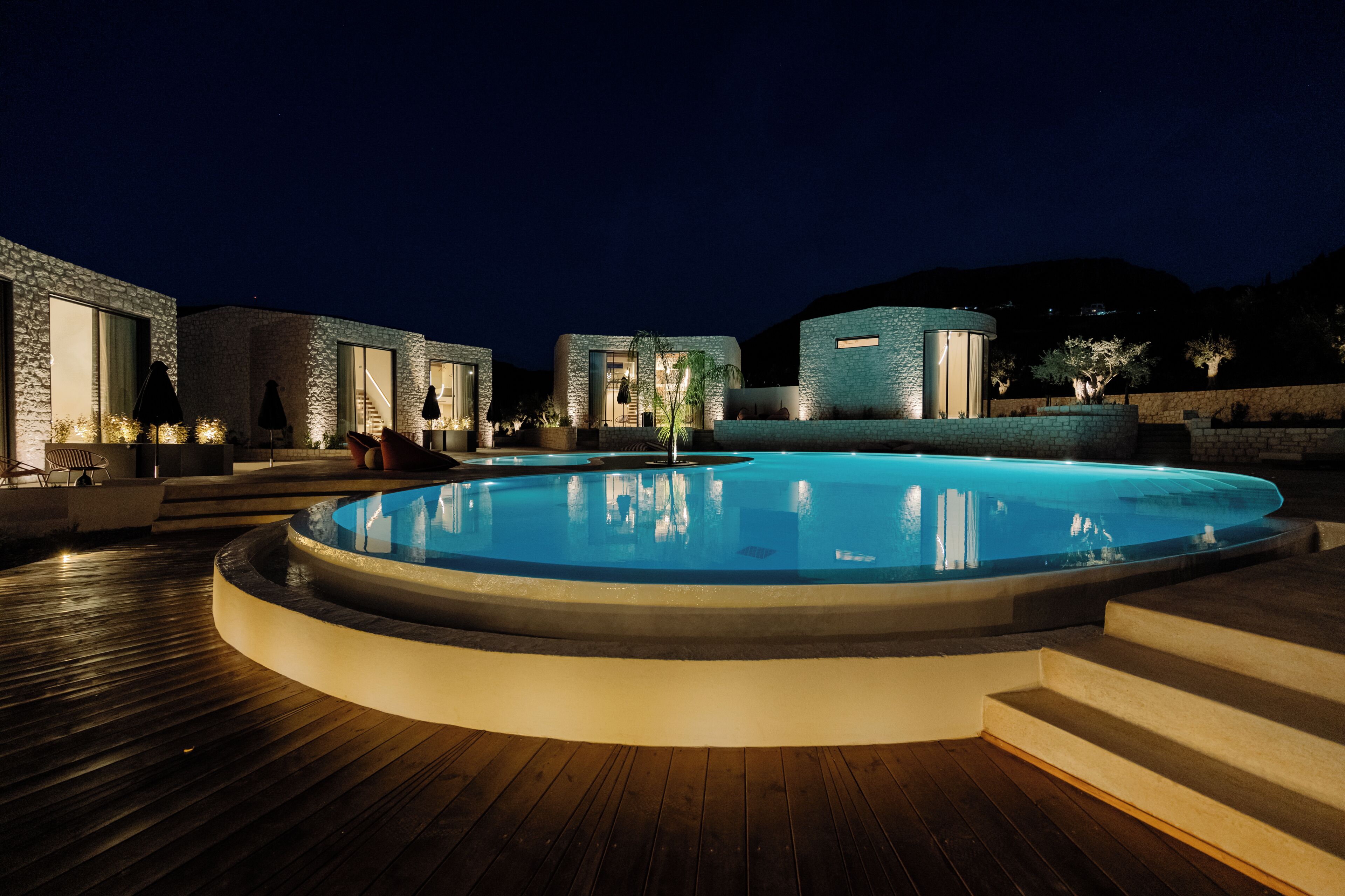 A swimming pool with a wooden deck, stone walls, and a night-time setting.