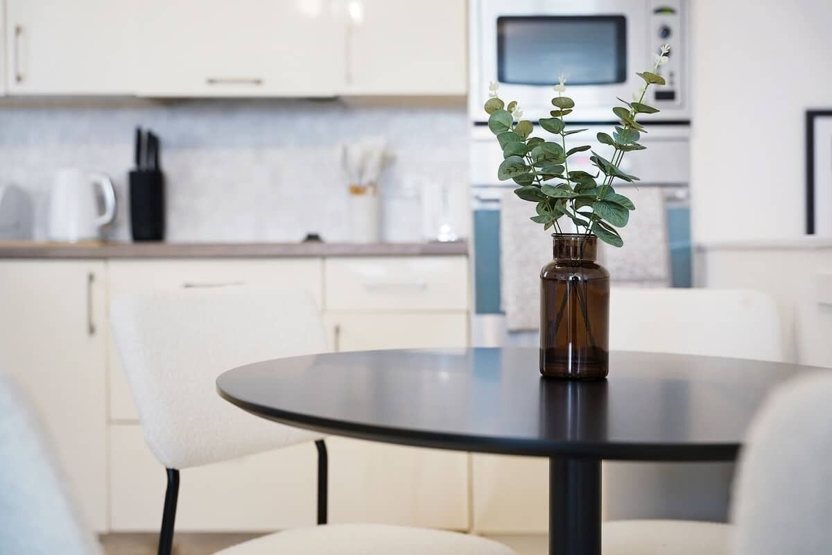 A modern kitchen with white cabinets, a microwave, and a vase with green plants on a round table.