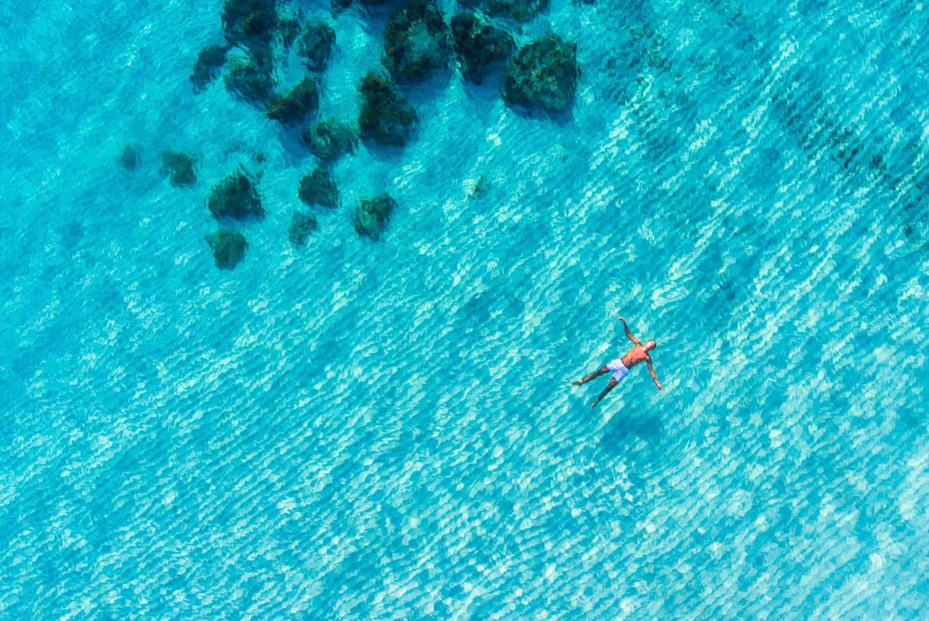 A person swimming in clear blue water with rocks in the background.