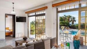 A modern living room with a sofa, a dining table with chairs, a TV mounted on the wall, and large windows with a view of a pool and palm trees.