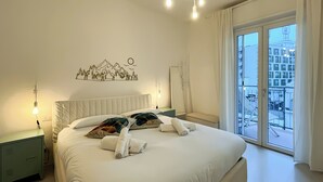 Ein Hotelzimmer mit Bett, Nachttisch, Wandlampe, Fenster mit Blick auf Gebäude und einer Berglandschaft an der Wand.
