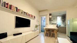Ein modernes Wohnzimmer mit Flachbildfernseher, Bücherregal, Essbereich mit Tisch und Stühlen sowie einer durchgehenden Tür erkennbaren Küche.