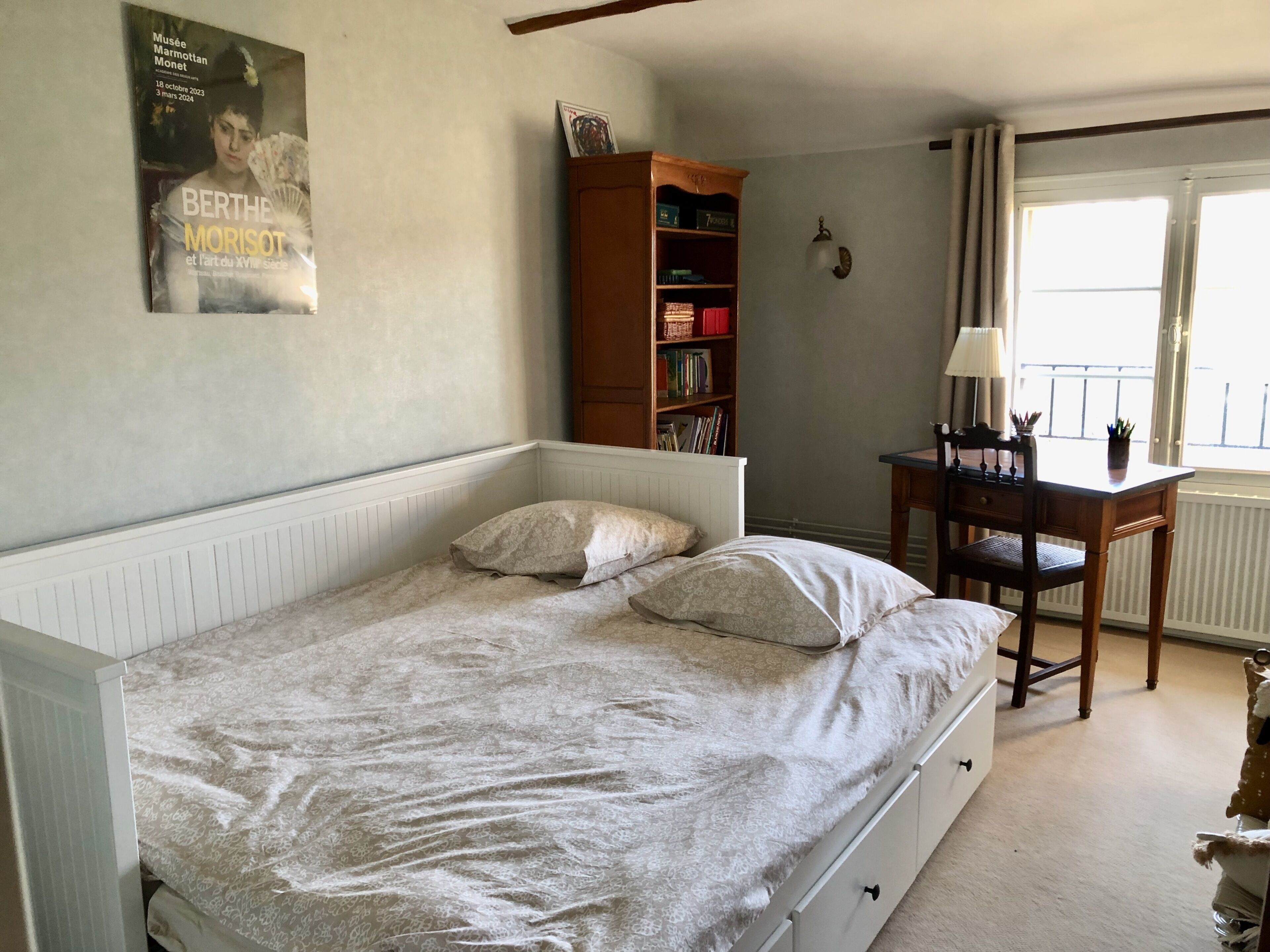 A bedroom with a single bed, a wooden bookshelf, a desk with a chair, and a window with curtains.