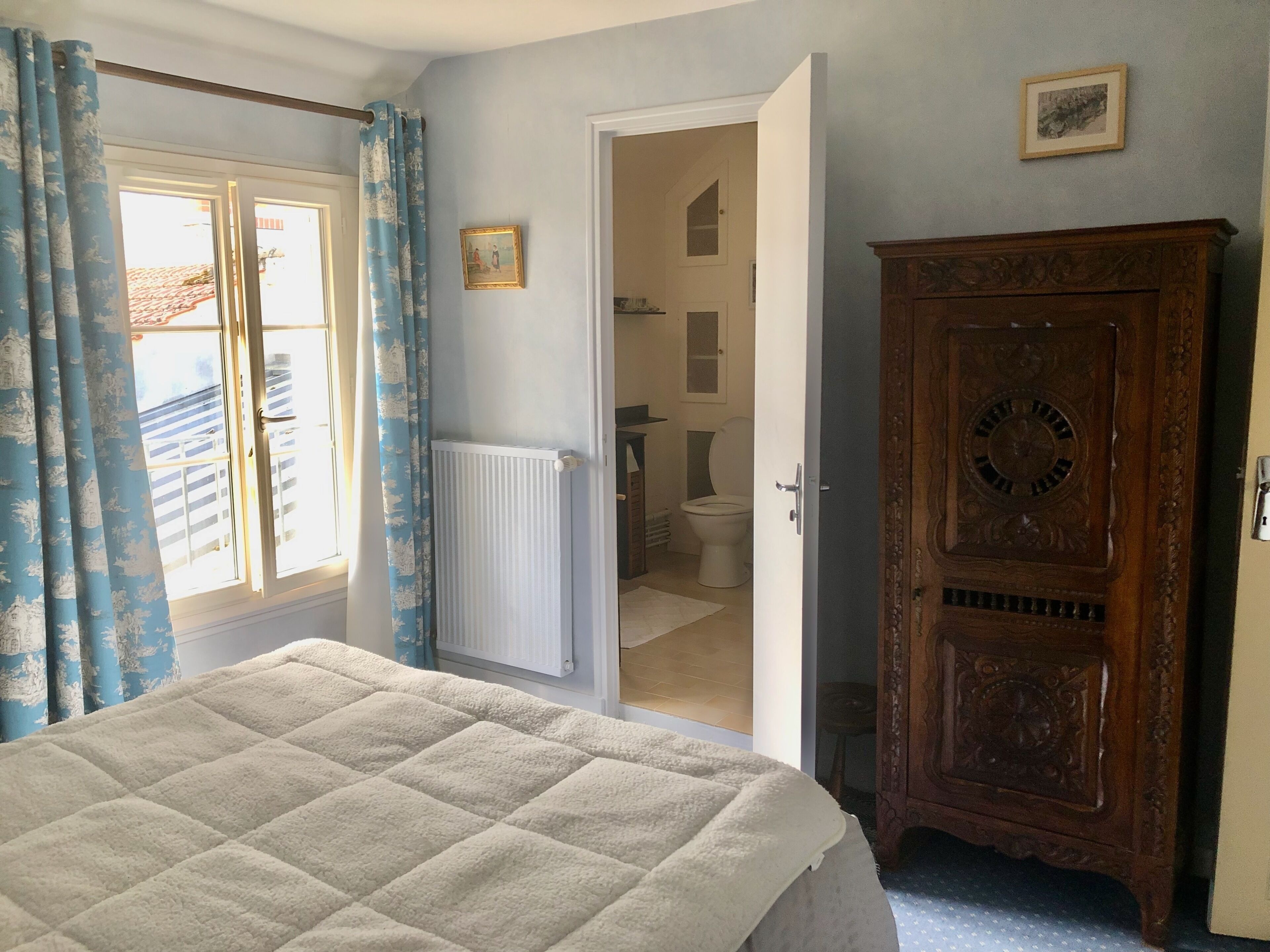 Ein Schlafzimmer mit einem Bett, einem Holzschrank, einem Heizkörper und einem durchgegangenen Türrahmen sichtbaren Badezimmer.