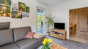 A modern living room with a grey sofa, a coffee table with flowers, a TV on a wooden stand, and a dining area with wooden furniture. - Apartment 'Burgquartier' with Balcony and Wi-Fi (Schmallenberg)