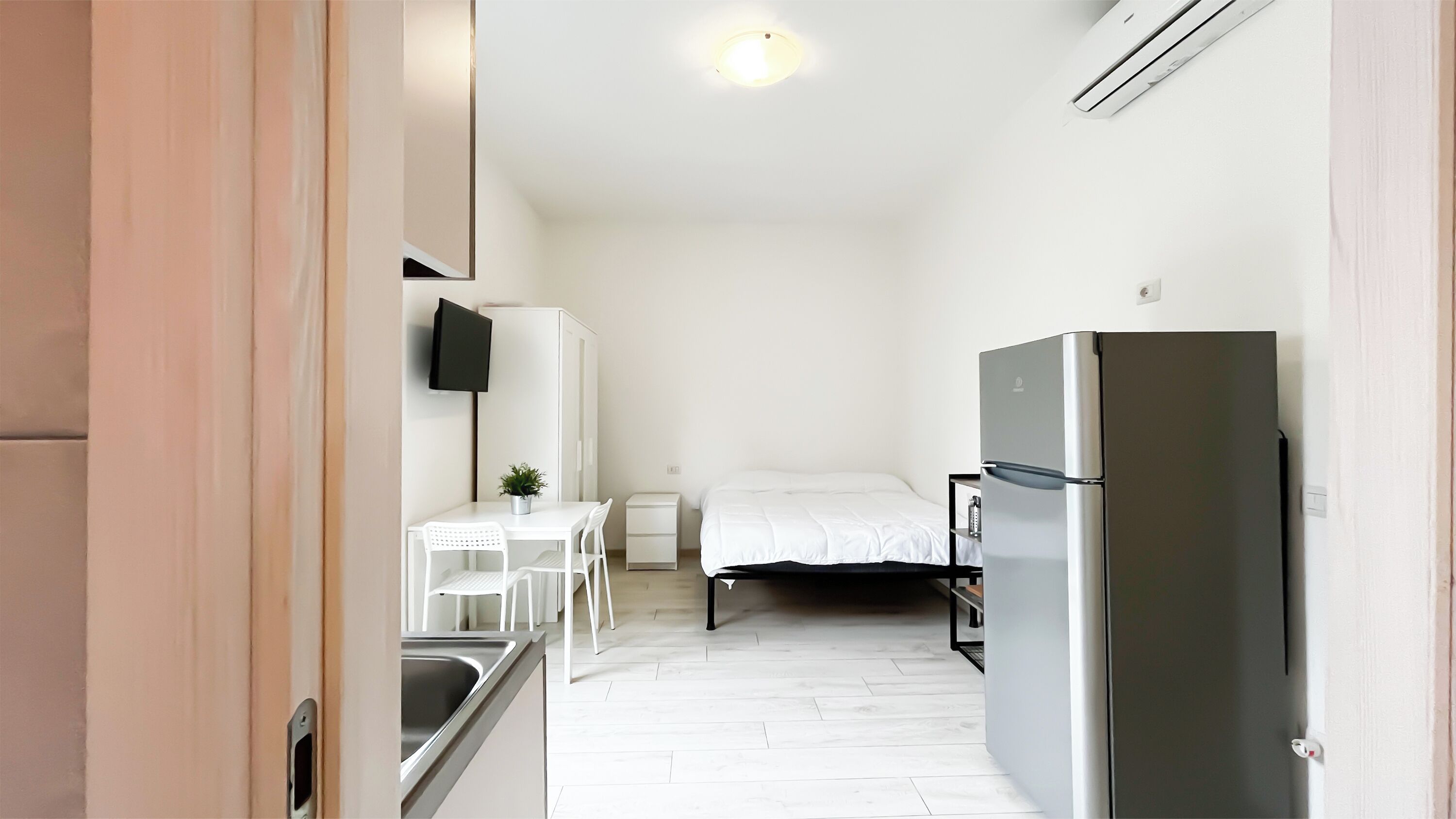 A compact kitchenette with a refrigerator, a small table with chairs, a TV mounted on the wall, and a bed with a white mattress.