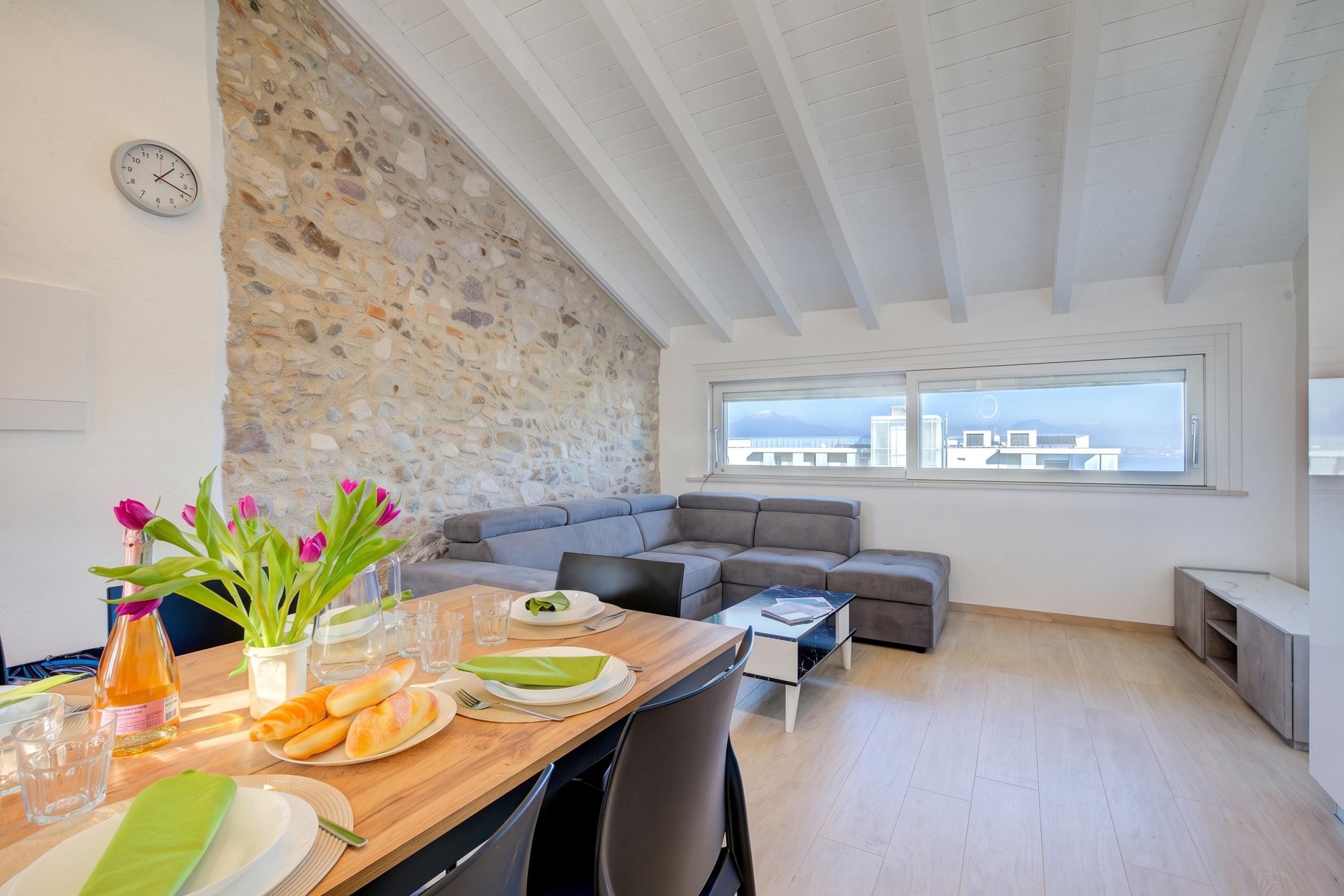 A modern living space with a stone wall, a dining area with a table set for two, a grey sofa, and a city view through large windows.