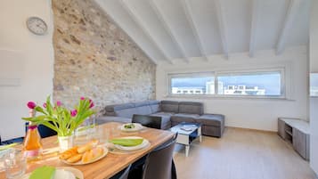 Ein moderner Wohnraum mit Steinwand, einem Essbereich mit Tisch für zwei Personen, einem grauen Sofa und Blick auf die Stadt durch große Fenster.