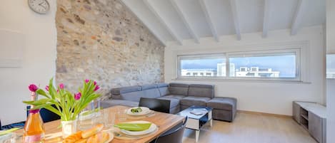 A modern living space with a stone wall, a dining area with a table set for two, a grey sofa, and a city view through large windows.
