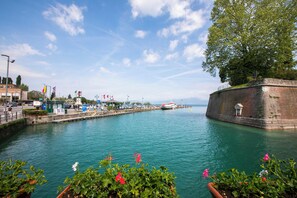 A waterfront view with a dock, a large tree, and a building with flags. - Peschiera Apartments Near The Lake (Peschiera del Garda)