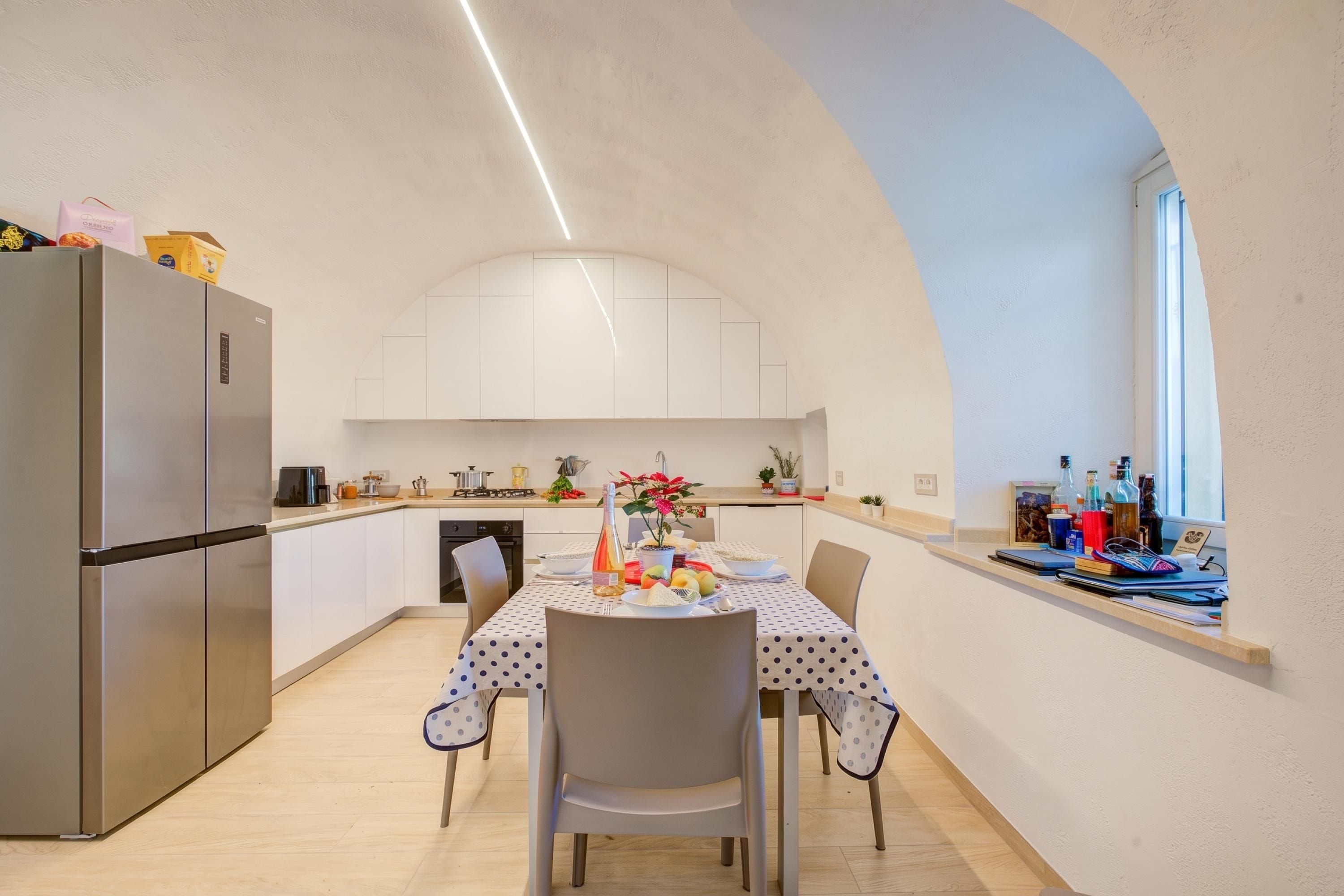 Cozinha moderna com mesa de jantar posta para dois, geladeira e janela com garrafas no balcão.
