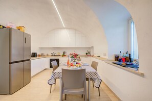 Eine moderne Küche mit Esstisch für zwei Personen, einem Kühlschrank und einem Fenster. Auf der Arbeitsplatte stehen Flaschen.