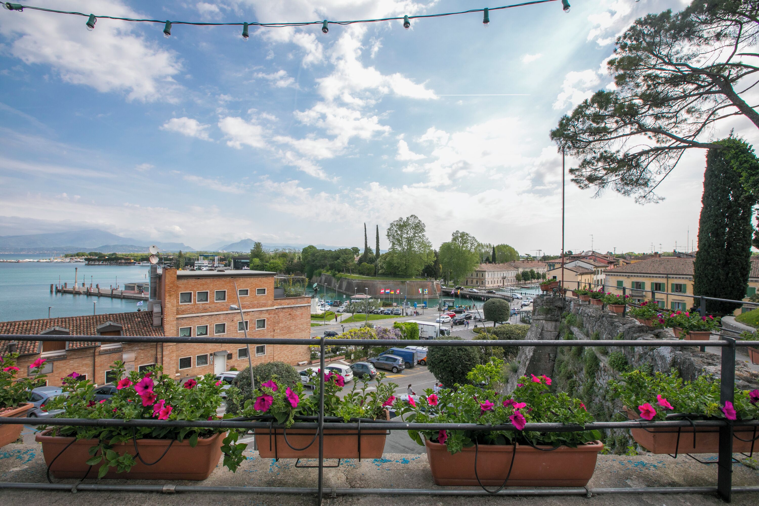 Un balcon agrémenté de jardinières en terre cuite remplies de fleurs, avec vue sur un port de plaisance, des bâtiments et un ciel dégagé.