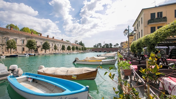 A waterfront area with boats docked, a row of buildings, and outdoor seating.
