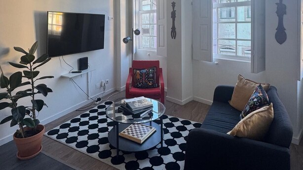 Una sala de estar con televisor, una silla roja, un sofá, una mesa de centro y un ventanal grande.