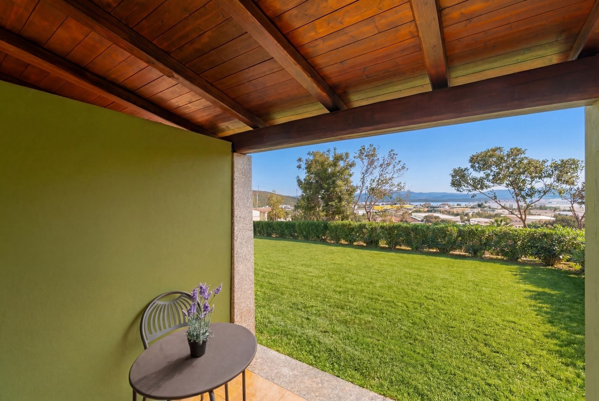 Una veranda con tetto in legno, un tavolo con una pianta in vaso e vista su un prato verde e un paesaggio in lontananza.