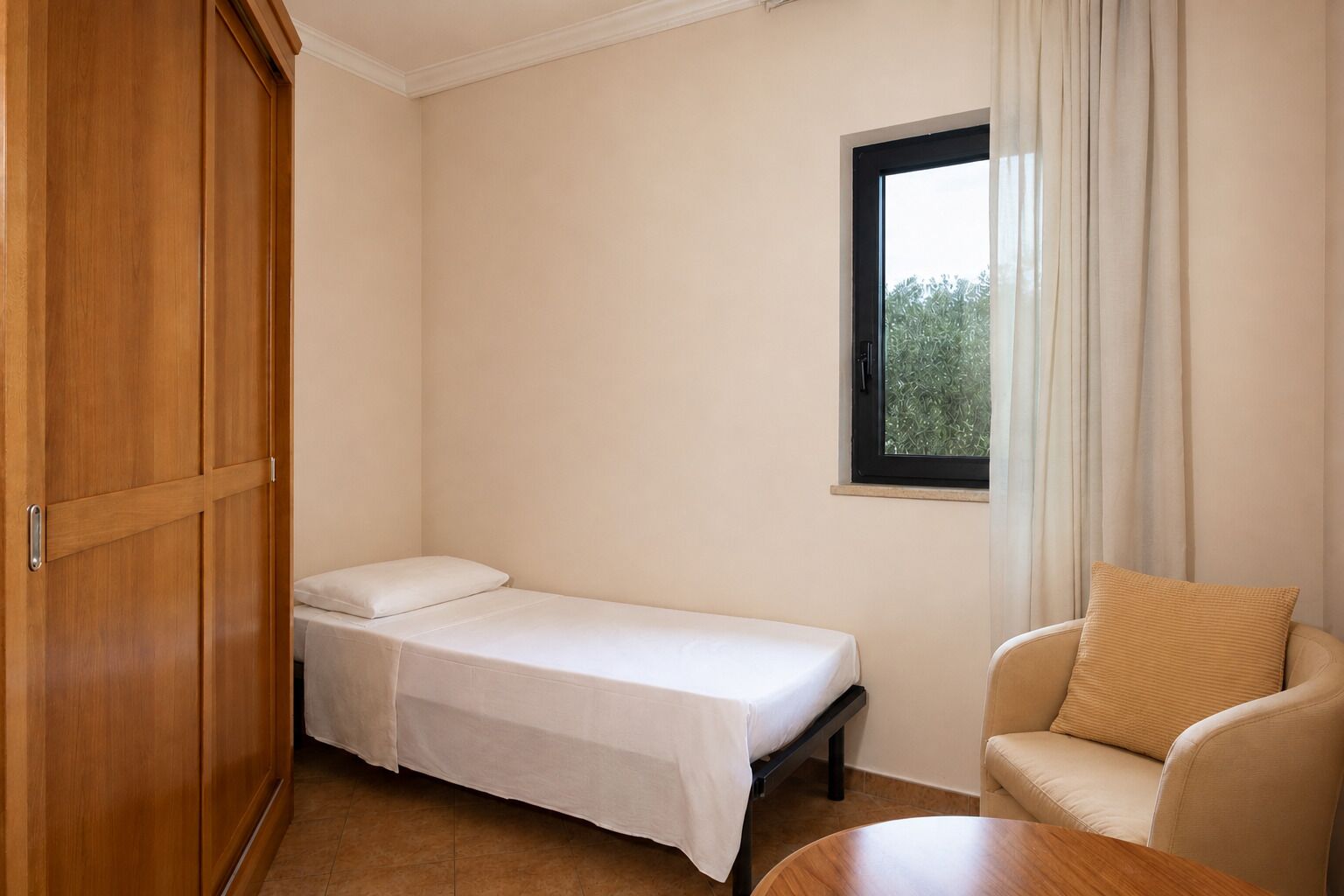 Una habitación pequeña con cama individual, un armario de madera, un ventanal con cortinas y un sofá color crema.