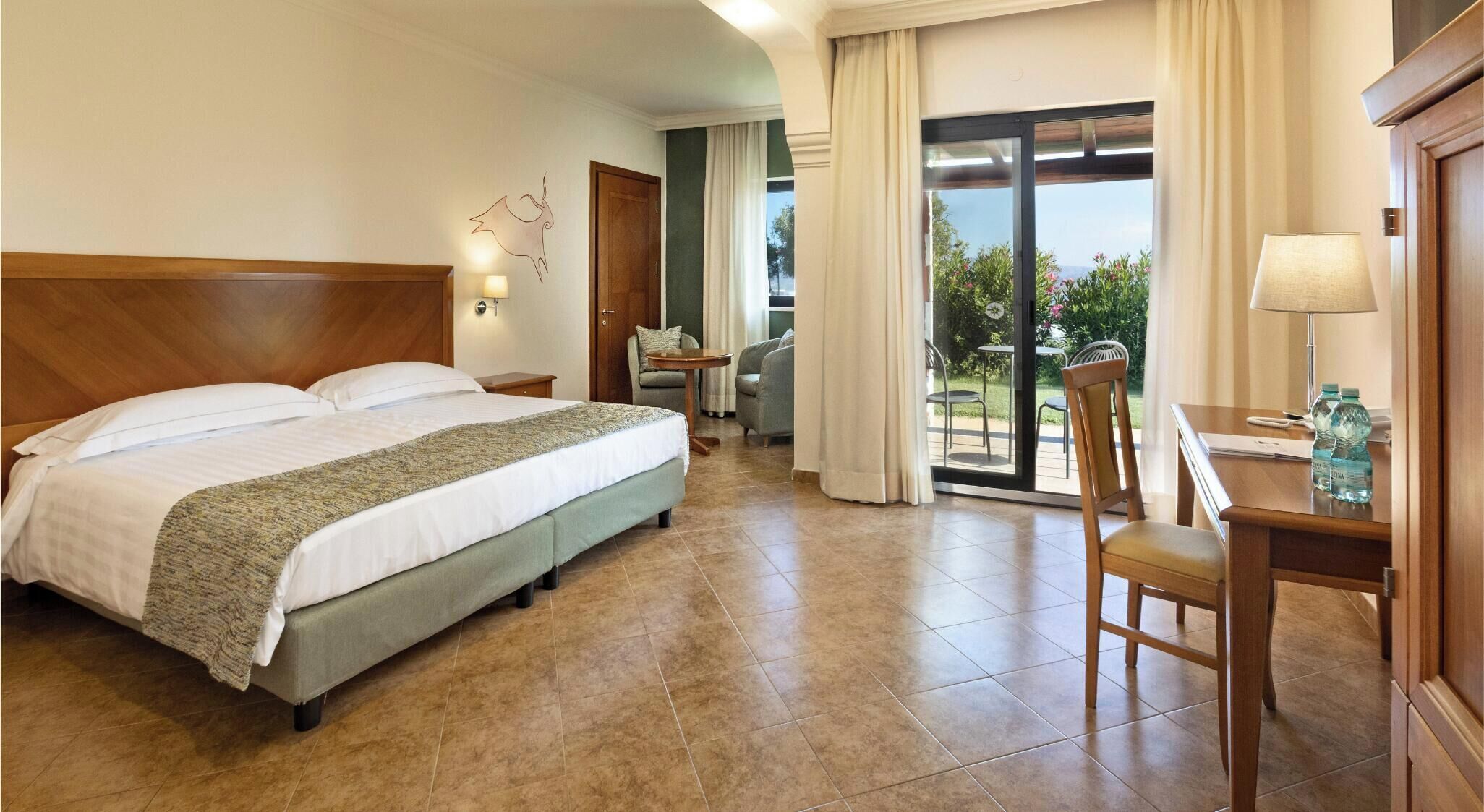 Ein Hotelzimmer mit einem großen Bett, einem Schreibtisch mit Stuhl, einem Tisch mit Lampe und Blick ins Freie durch eine Glastür.