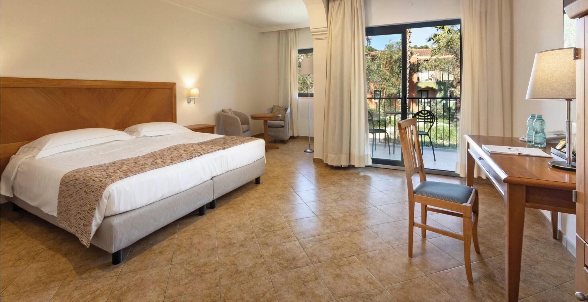 A hotel room with a large bed, a desk, a chair, and a view of the outdoors through a sliding door.