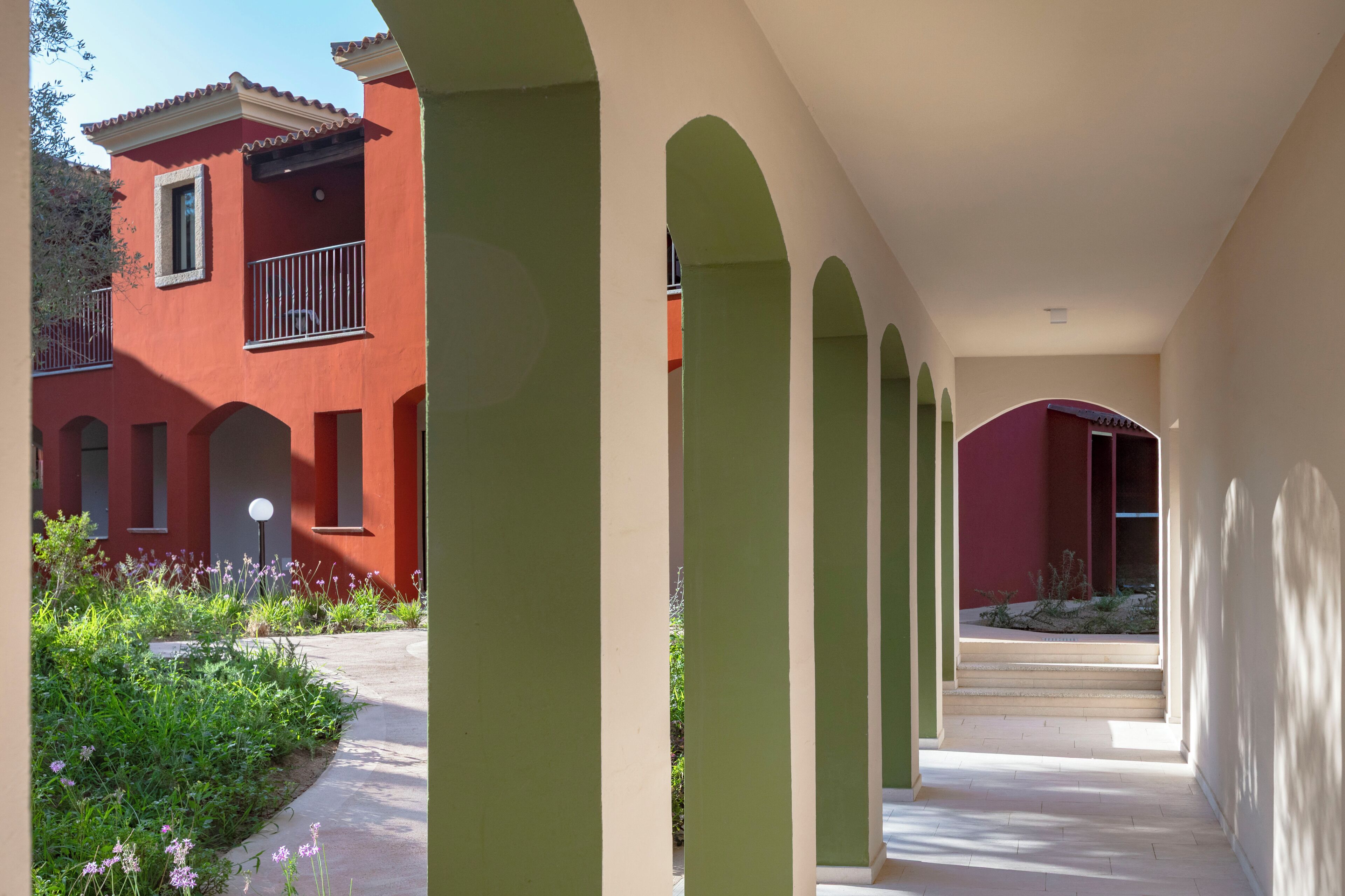 A corridor with arches, a red building, and a garden with purple flowers.