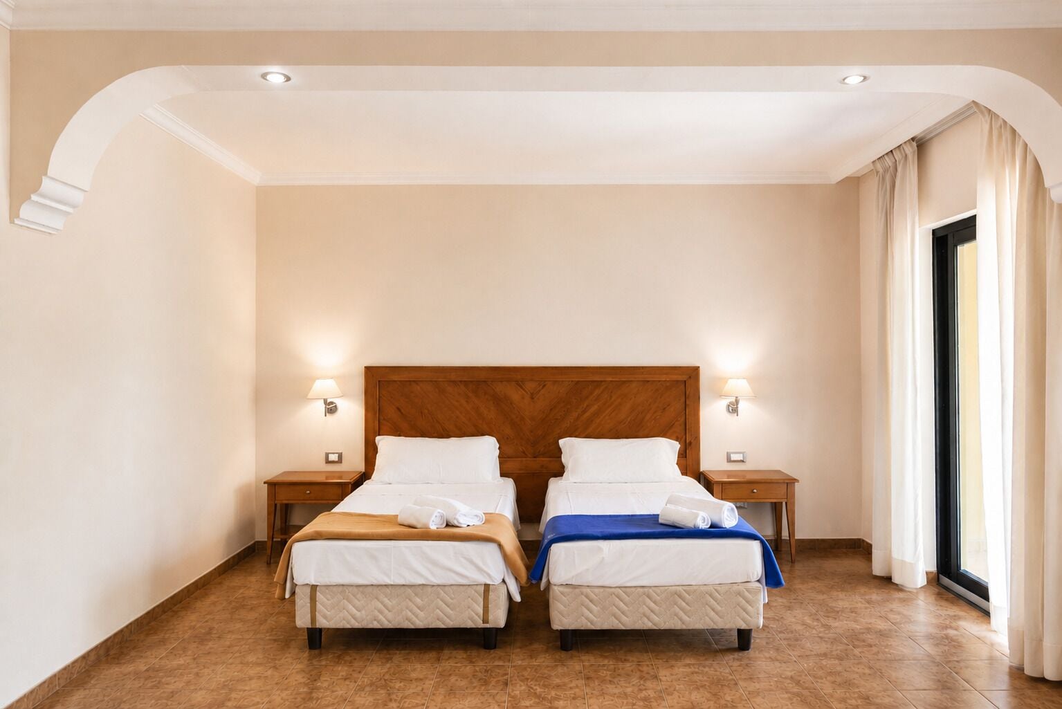 Une chambre d’hôtel avec deux lits, une tête de lit en bois, des lampes de chevet et une grande fenêtre avec des rideaux.