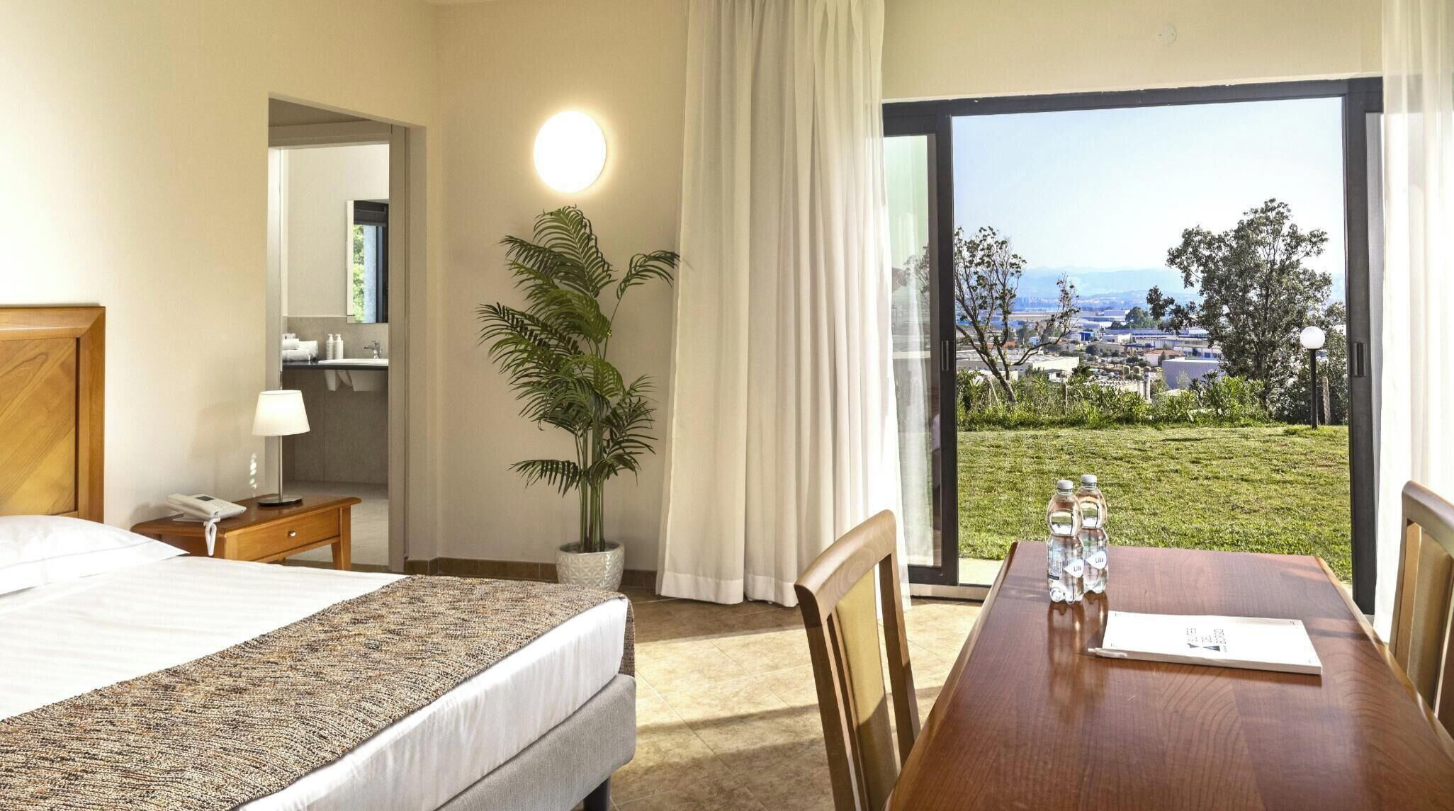 A hotel room with a bed, a desk with a chair, a potted plant, a bathroom with a mirror, and a view of the outdoors.