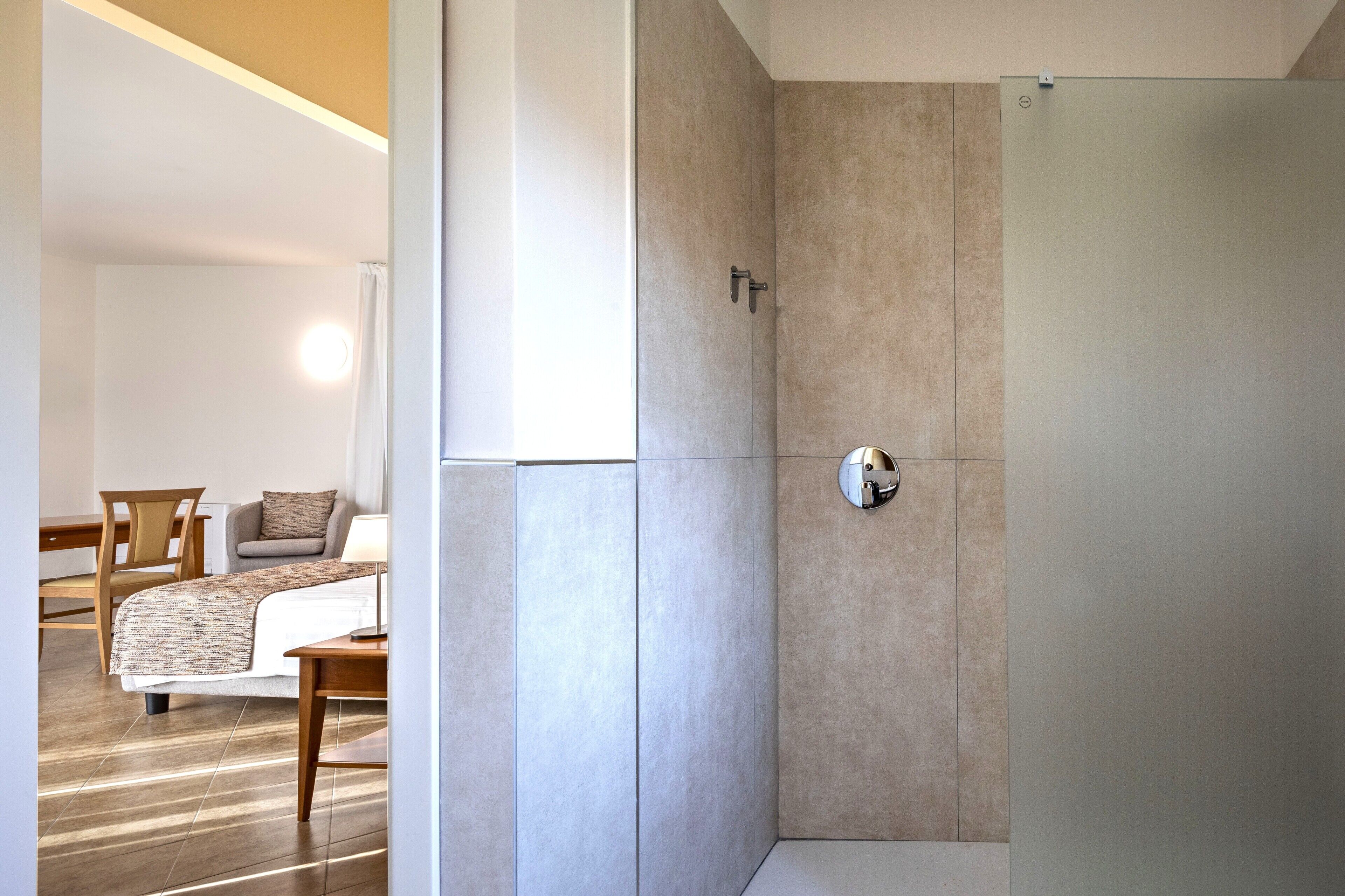 A modern bathroom with a shower, a toilet, and a sink. There is a wooden chair and a small table with a lamp in the adjacent room.