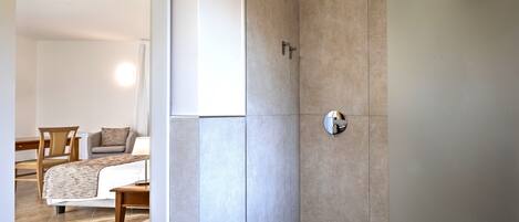 A modern bathroom with a shower, a toilet, and a sink. There is a wooden chair and a small table with a lamp in the adjacent room.
