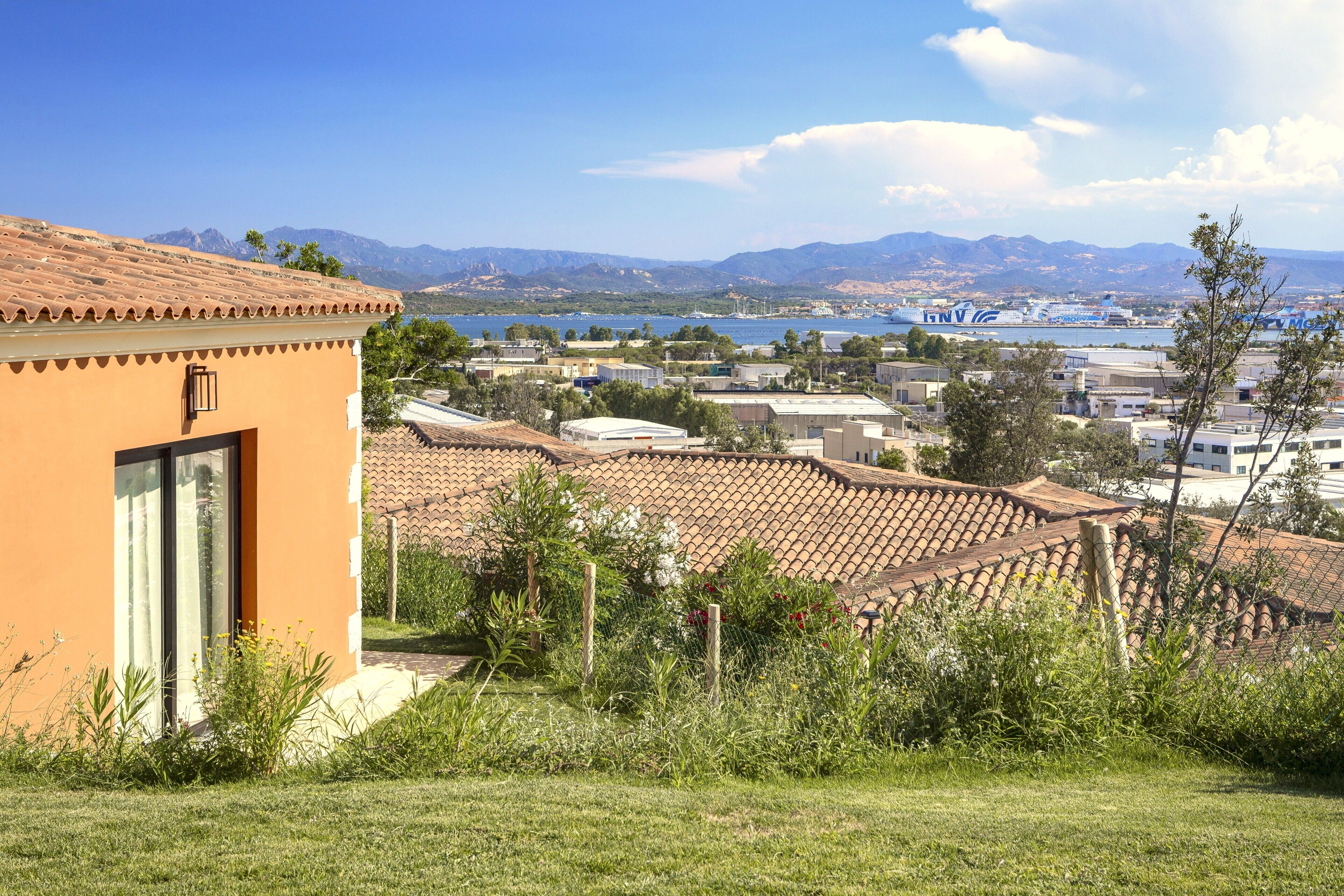 A coastal town with terracotta-roofed buildings, a body of water, and mountains in the background.