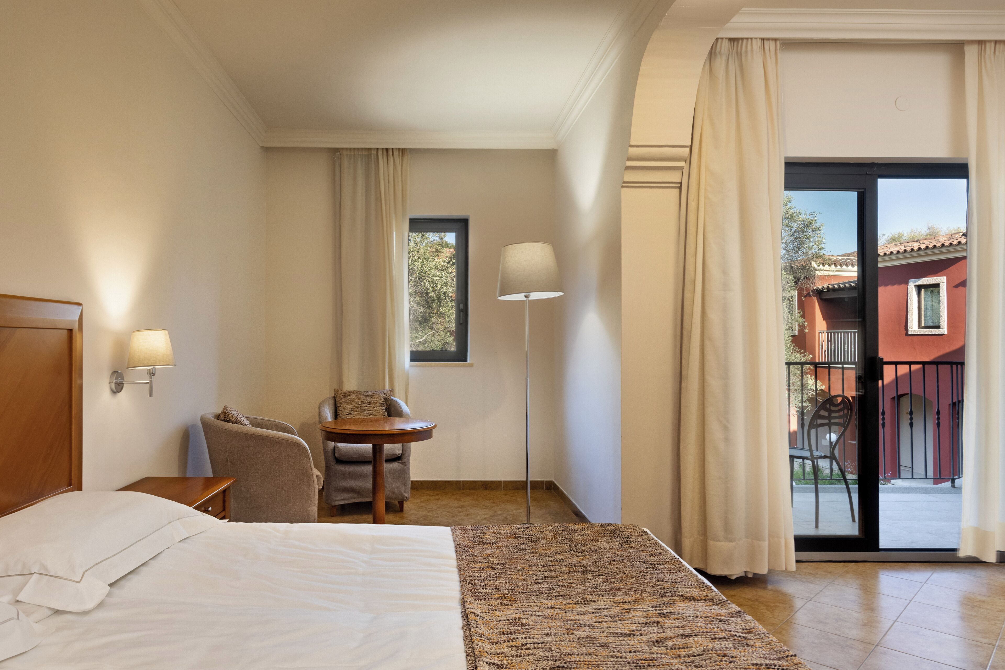 A hotel room with a bed, a chair, a small table, a lamp, and a view of the outside.