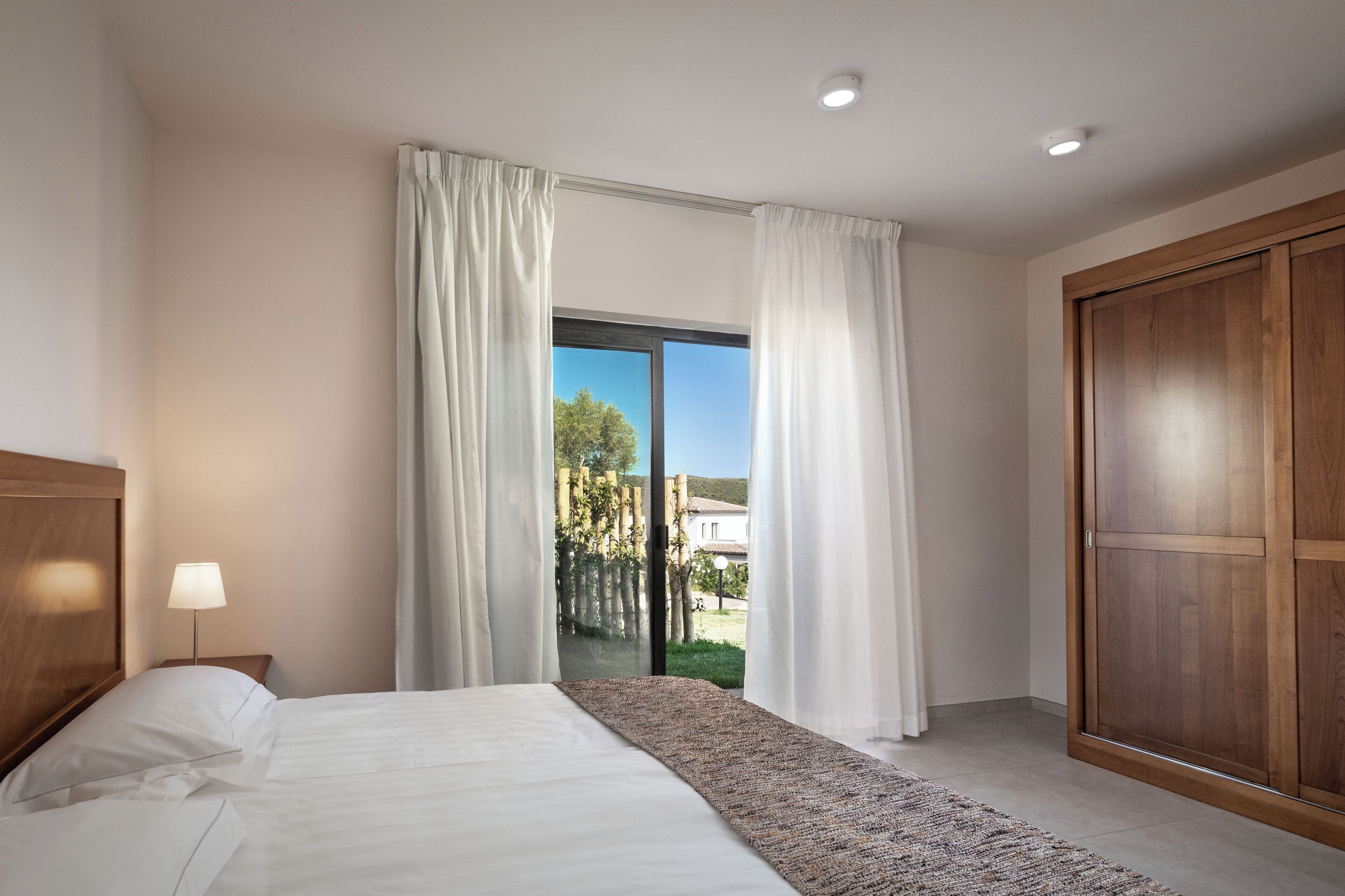 Habitación de hotel con cama, mesita de noche, lámpara, ventana grande con cortinas y armario de madera.