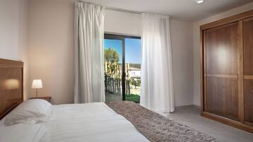 A hotel room with a bed, a nightstand, a lamp, a large window with curtains, and a wooden wardrobe.