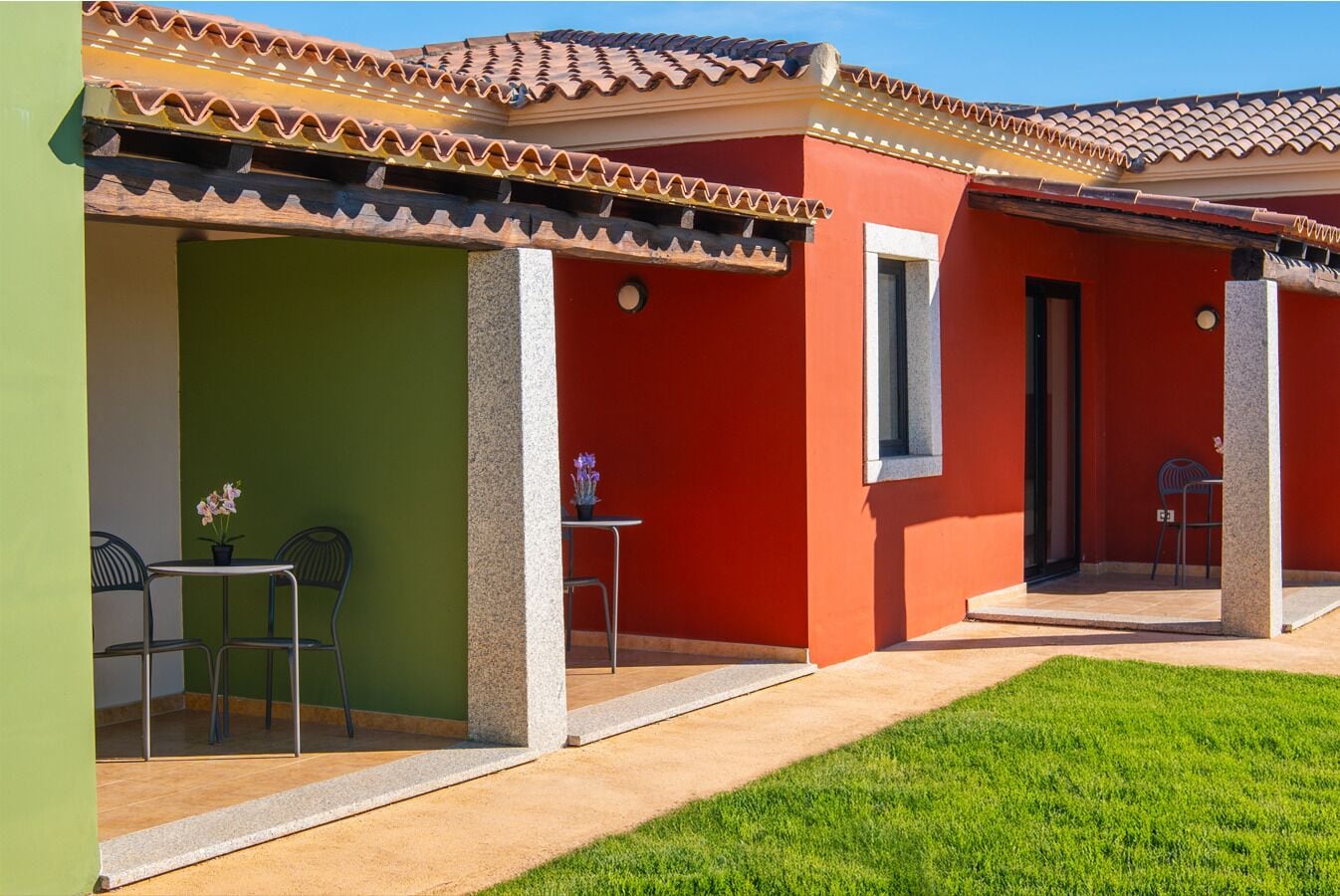 A building with a tiled roof, a green wall, and a red wall with a window. There are two outdoor tables with chairs and a potted plant.